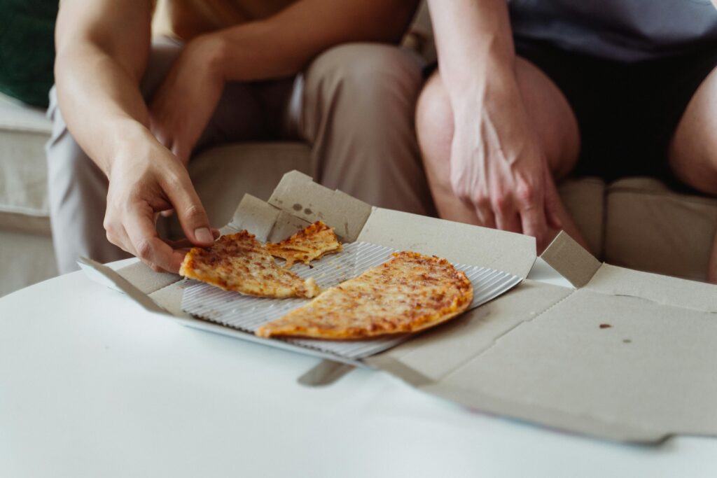 Two friends sharing a pizza from a takeaway box during a casual indoor meal.