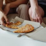 Two friends sharing a pizza from a takeaway box during a casual indoor meal.