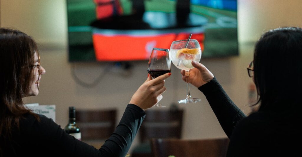 Two women clinking wine glasses at a restaurant with a sports broadcast on TV in the background.