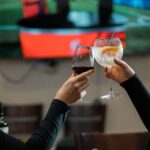 Two women clinking wine glasses at a restaurant with a sports broadcast on TV in the background.